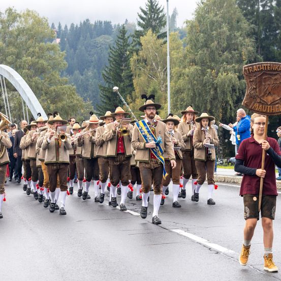 Eine Blaskapelle in traditioneller bayerischer Tracht tritt im Freien auf, mit einem Banner, das 'Trachtenka' im Vordergrund zeigt. Bäume und eine Brücke sind im Hintergrund zu sehen.