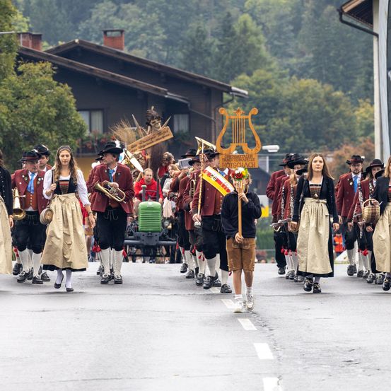 Eine Blaskapelle in traditioneller bayerischer Kleidung, mit Musikinstrumenten, geht eine Straße entlang. Hinter ihnen sind Häuser und Bäume sichtbar.
