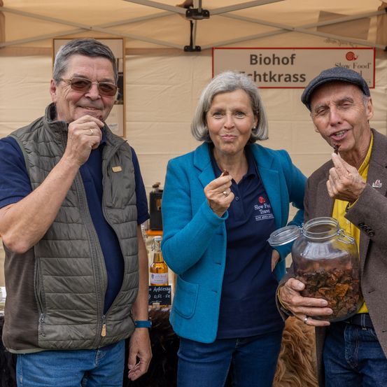 Drei Personen stehen unter einem Zelt, lächeln und essen etwas. Der Mann links trägt eine Weste und eine Brille. Die Frau in der Mitte trägt eine blaue Jacke und hat kurze Haare. Der Mann rechts hält ein Glas.