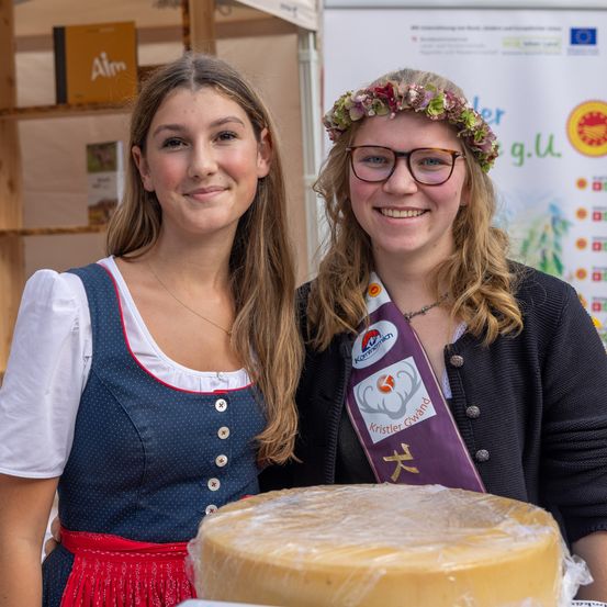 Zwei Frauen stehen neben einem großen runden Käse, eine in traditioneller bayerischer Tracht, die andere mit Blumenkranz und Brille. Dahinter befindet sich ein Werbebanner.