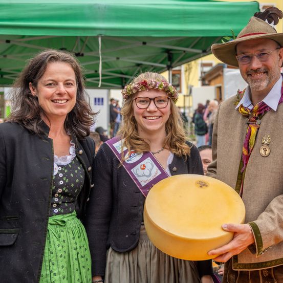 Drei Menschen stehen lächelnd unter einem grünen Zelt. Eine Frau links, eine Frau in der Mitte mit einem Blumenkranz und ein Mann rechts, der einen großen gelben Käse hält.