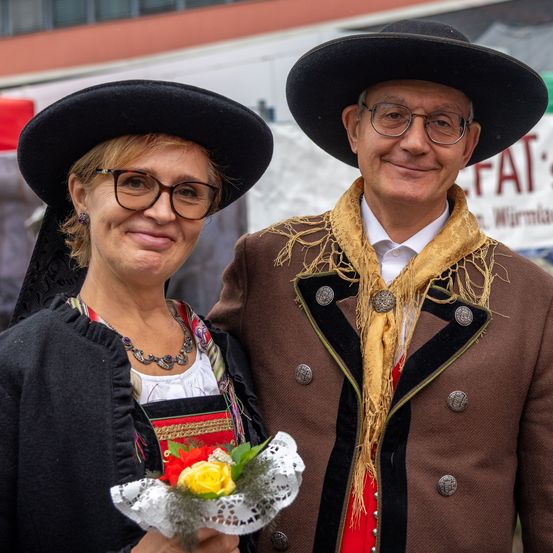 Ein lächelndes Paar in traditioneller Kleidung steht eng beieinander, wobei die Frau einen Blumenstrauß hält. Der Mann trägt einen braunen Mantel mit gelbem Besatz und einen Schal. Sie tragen beide breitkrempige Hüte.
