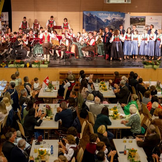 Eine Musikband tritt auf der Bühne vor einem sitzenden Publikum auf, einige klatschen. Die Bühne ist mit Blumen und einer Schweizer Flagge geschmückt. Im Zuschauerbereich sind Tische aufgestellt.