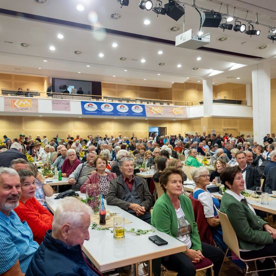 Ein großer Saal ist mit Menschen gefüllt, die an Tischen sitzen, wahrscheinlich für eine Veranstaltung. Der Raum hat Banner und Lichter. Die Teilnehmer sind vielfältig und tragen verschiedene Kleidungsstücke. Es sind viele Menschen versammelt, was eine lebhafte Atmosphäre schafft.