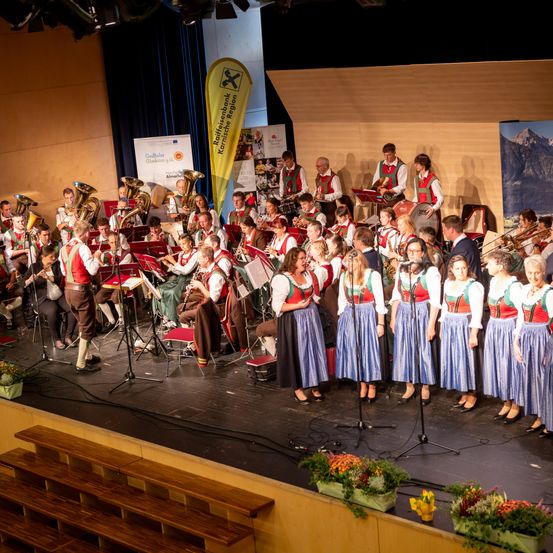 Ein Orchester auf der Bühne, mit Mitgliedern in traditioneller Kleidung, tritt in einem Saal mit einer Gebirgskulisse und einem Banner hinter ihnen auf.