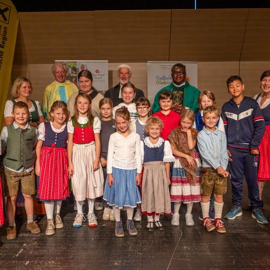 Eine Gruppe von Kindern und Erwachsenen in traditioneller bayerischer Kleidung steht lächelnd auf der Bühne. Ein Priester und Erwachsene sind ebenfalls anwesend. Hinter ihnen befinden sich Banner, eines mit dem Text 'Slow Food Travel'.