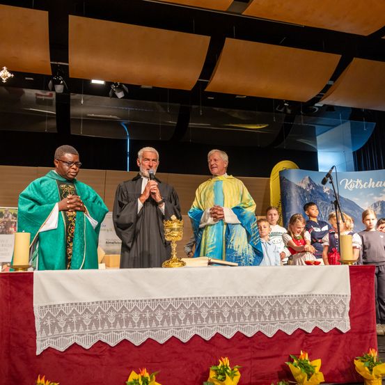 Drei Priester stehen auf einer Bühne, einer hält ein Mikrofon. Dahinter beobachten Kinder. Altar und Kerzen schmücken die Bühne.