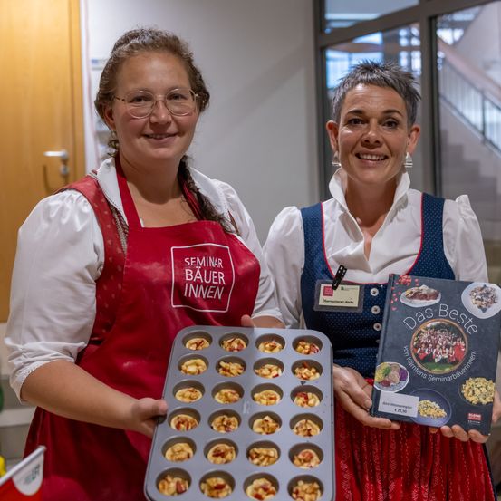 Zwei Frauen in traditioneller Kleidung halten ein Tablett mit Muffins und ein Kochbuch. Die Frau auf der linken Seite trägt ein rotes Schürzen mit Text. Die Frau auf der rechten Seite hält ein Kochbuch und lächelt. Hinter ihnen ist eine weiße Wand mit einer Tür.