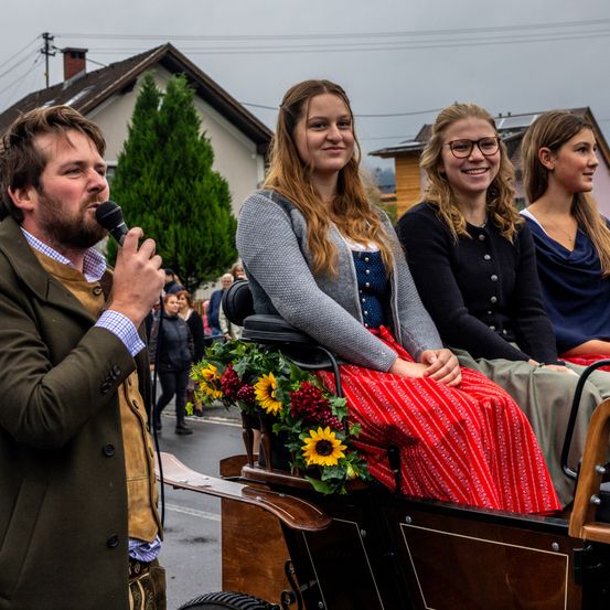 Ein Mann in einem Anzug spricht in ein Mikrofon, während drei Frauen in traditioneller Kleidung auf einer Kutsche sitzen und lächeln. Die Kutsche ist mit Blumen geschmückt, und Menschen laufen dahinter.