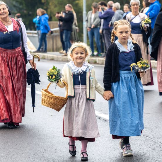 Zwei junge Mädchen, eines mit einem Korb und das andere mit Blumen, gehen gemeinsam auf einer Straße. Dahinter beobachten mehrere Menschen, einige mit Kameras, die Prozession.