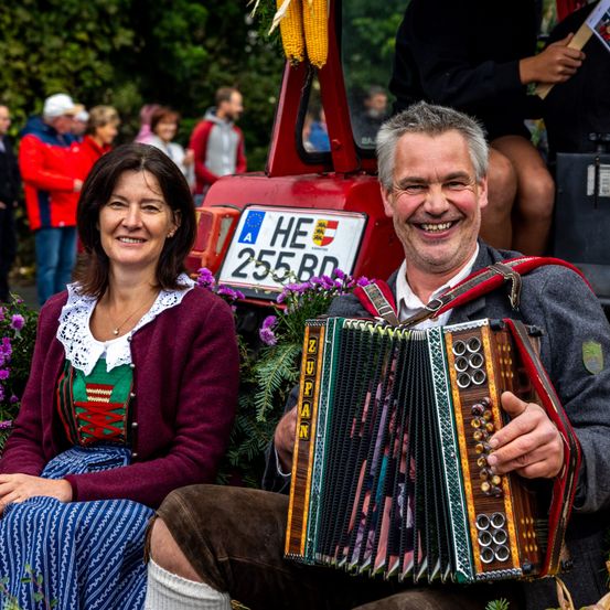 Ein lächelnder Mann und eine Frau posieren vor einem roten Traktor, mit einer Menschenmenge im Hintergrund, an einem sonnigen Tag.