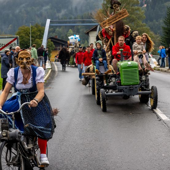 Eine Frau in einem Kostüm fährt Fahrrad, während ein Traktor eine Familie trägt und hinter ihr auf der Straße fährt. Menschen gehen am Straßenrand.
