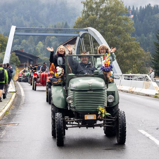 Eine Gruppe von Menschen fährt auf grünen Traktoren, die mit Blumen geschmückt sind, eine Straße hinunter mit einer Brücke im Hintergrund.