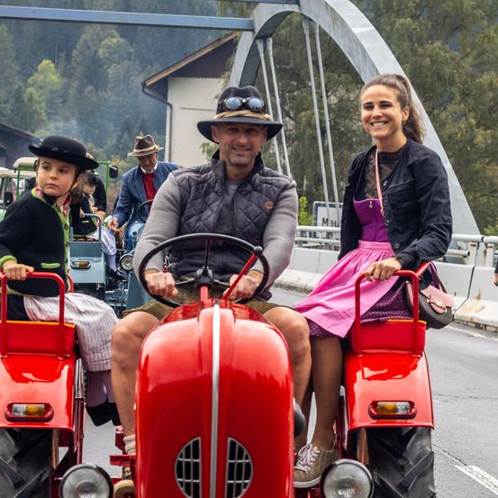 Drei Personen fahren auf roten Traktoren auf einer Straße, mit einer Brücke und Bäumen im Hintergrund.