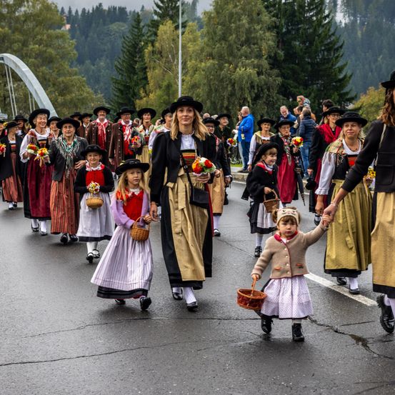 Eine Parade von Menschen in traditionellen Kostümen, einige mit Blumen, geht unter einer Brücke über eine Straße. Ein kleines Kind hält die Hand einer Erwachsenen vor sich.