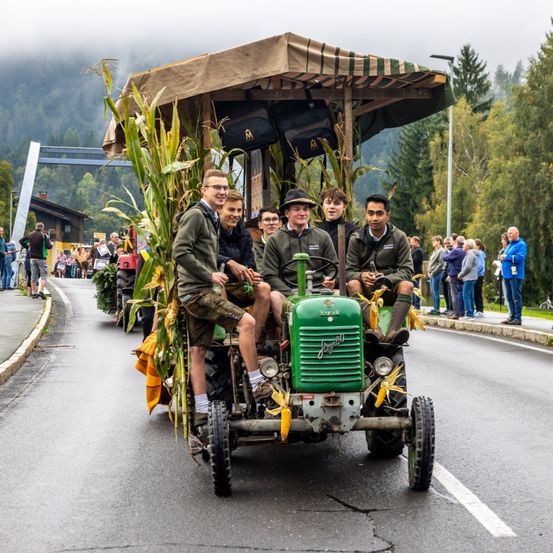 Fünf junge Männer sitzen auf einem grünen Oldtimer-Traktor mit Maisdach und fahren auf einer Straße, wo Menschen am Bürgersteig zusehen.