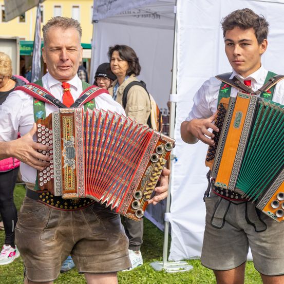 Zwei Männer in traditioneller bayerischer Kleidung spielen Akkordeons in einer Freiluftkulisse. Ein Mann ist älter und der andere jünger. Sie stehen auf einem Rasenbereich mit einem weißen Zelt im Hintergrund.