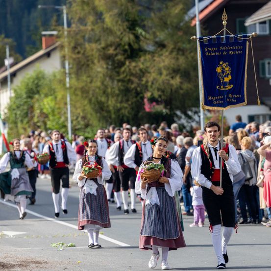 Eine Gruppe von Menschen in traditionellen Kostümen zieht mit Blumenkörben die Straße entlang. Zuschauer beobachten von der Straßenseite aus, und ein Mann hält eine Flagge vorne.