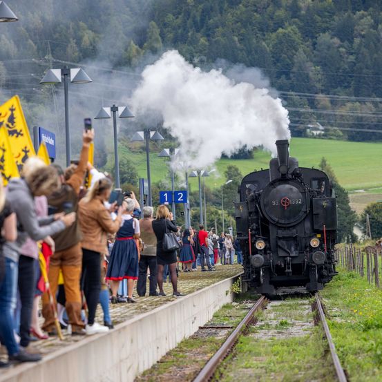 Eine schwarze Dampflokomotive auf den Gleisen, mit Menschen auf dem Bahnsteig. Der Zug stößt weißen Rauch aus und die Menschen machen Fotos.