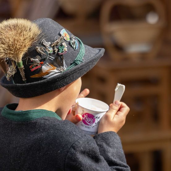 Ein Junge, der einen traditionellen Hut mit einer Feder und Abzeichen trägt, hält einen Becher und isst Eiscreme.