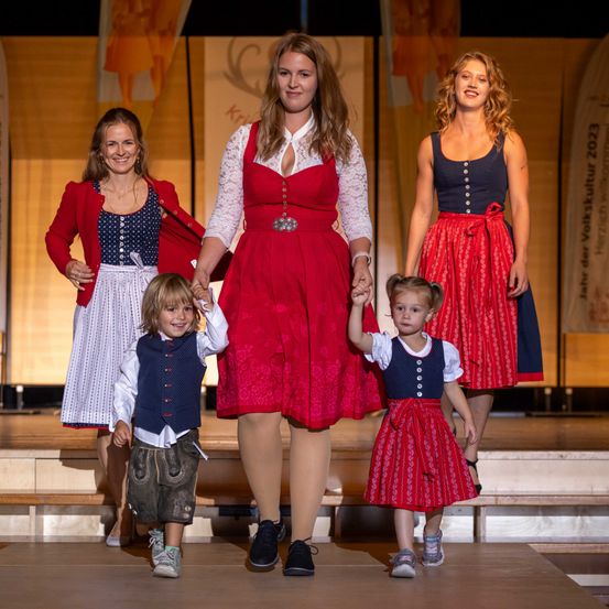 Vier Frauen in traditioneller bayerischer Kleidung und zwei Kinder stehen lächelnd auf der Bühne und halten Händchen. Die Frau in der Mitte trägt ein rotes Kleid, während die anderen rote und weiße Kleider tragen. Die Kinder sind in traditioneller Kleidung gekleidet.