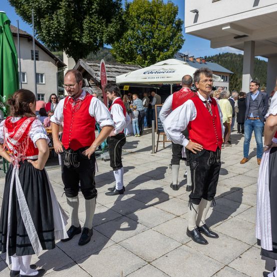 Mehrere Menschen in traditioneller Kleidung stehen auf einem Außenbereich, möglicherweise bei einem Festival. Ein Mann mit Brille schaut zur Seite. Hinter ihnen steht ein Zelt mit einem Schild, auf dem Edelweiss steht.