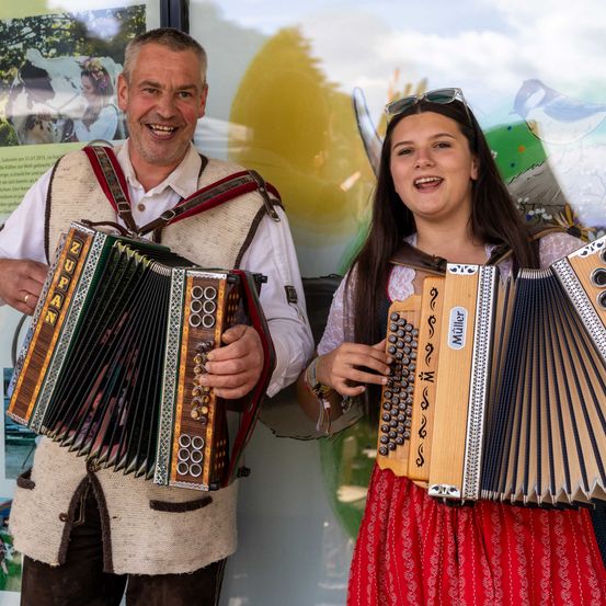 Ein Mann und eine Frau lächeln und spielen Akkordeons. Der Mann trägt eine traditionelle Kleidung. Sie stehen vor einer farbenfrohen Wandmalerei.