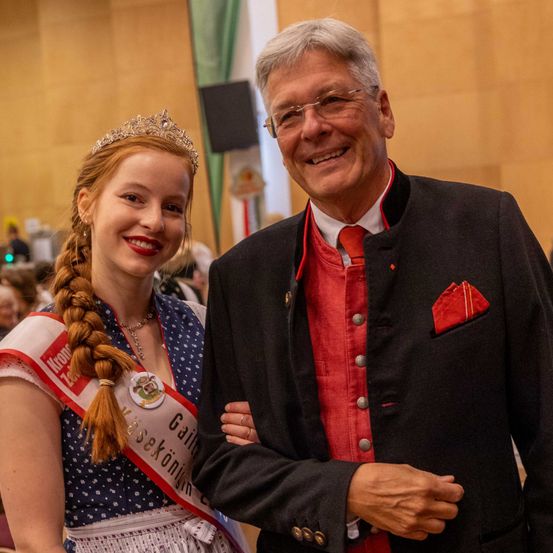 Ein Mann und eine Frau lächeln in einer festlichen Umgebung. Die Frau trägt eine Krone und eine Schärpe, die 'Miss' steht. Der Mann ist in einem roten Hemd, einem schwarzen Mantel und einer Brille gekleidet.