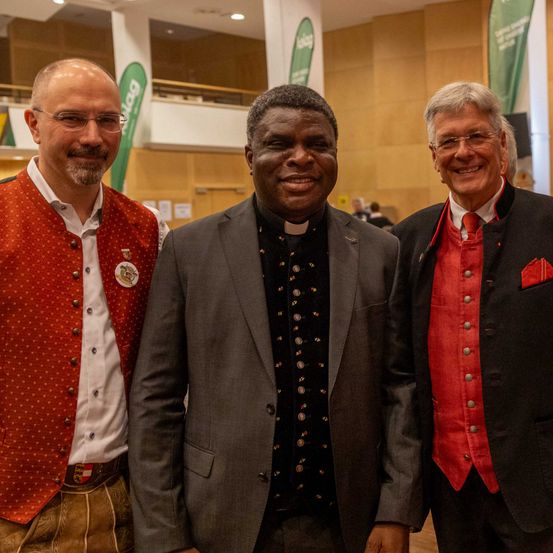 Drei Männer stehen zusammen und lächeln für die Kamera. Der Mann auf der linken Seite trägt eine rote Weste mit weißen Punkten. Der Mann in der Mitte, ein Priester, trägt eine schwarze Weste mit Knöpfen und einen weißen Kragen. Der Mann auf der rechten Seite trägt einen schwarzen Anzug und eine rote Weste.
