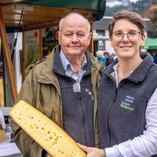 Ein Mann und eine Frau stehen nebeneinander und halten ein großes Stück Käse. Der Mann trägt eine Jacke und eine Brille, und die Frau trägt eine Brille. Sie lächeln und posieren für ein Foto. Hinter ihnen stehen mehrere Leute, und ein grünes Zelt ist auf der rechten Seite. In der Ferne gibt es Berge.