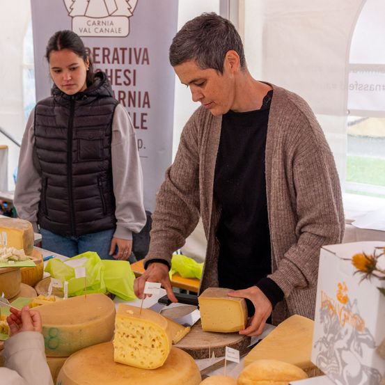 Ein Mann in einem braunen Strickcardigan schneidet Käse in einem Marktstand, eine Frau steht neben ihm. Dahinter steht ein Schild mit der Aufschrift 'Carnia Val Canale'.
