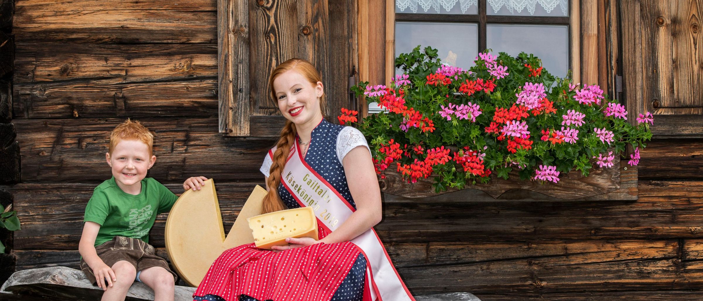 Eine Frau in traditioneller Kleidung hält ein Käsestück, während sie neben einem Jungen auf einer Bank vor einem Holzhaus mit Blumen sitzt.