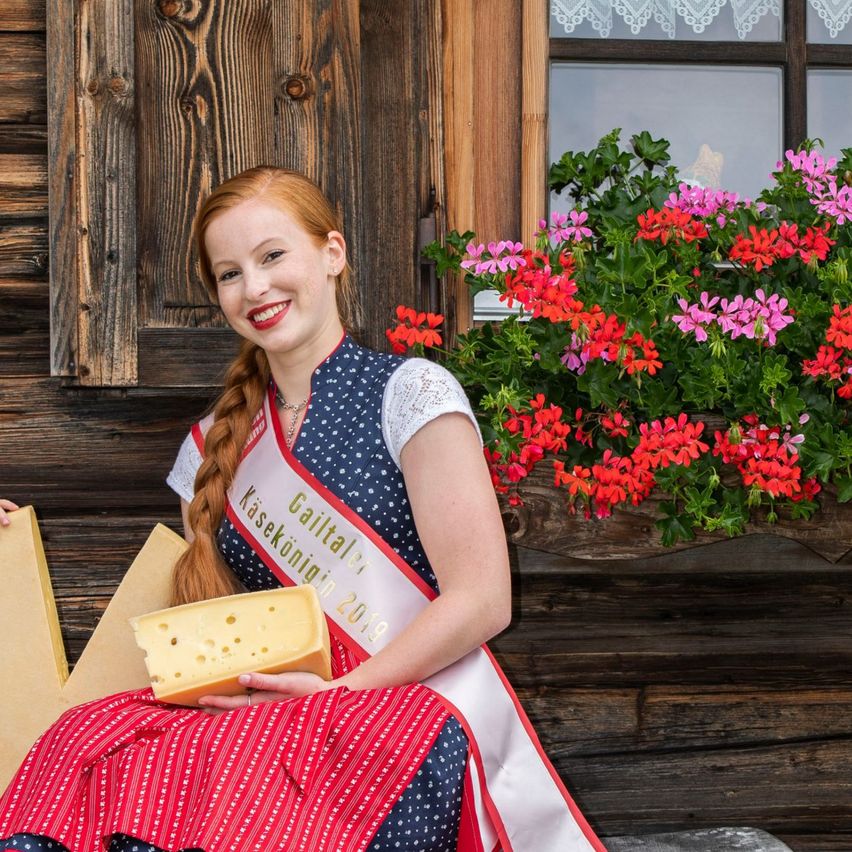 Eine Frau in traditioneller Kleidung hält ein Käsestück, während sie neben einem Jungen auf einer Bank vor einem Holzhaus mit Blumen sitzt.