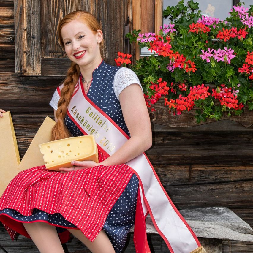 A woman in traditional costume holds a cheese slice while sitting beside a young boy on a bench in front of a wooden house with flowers.
