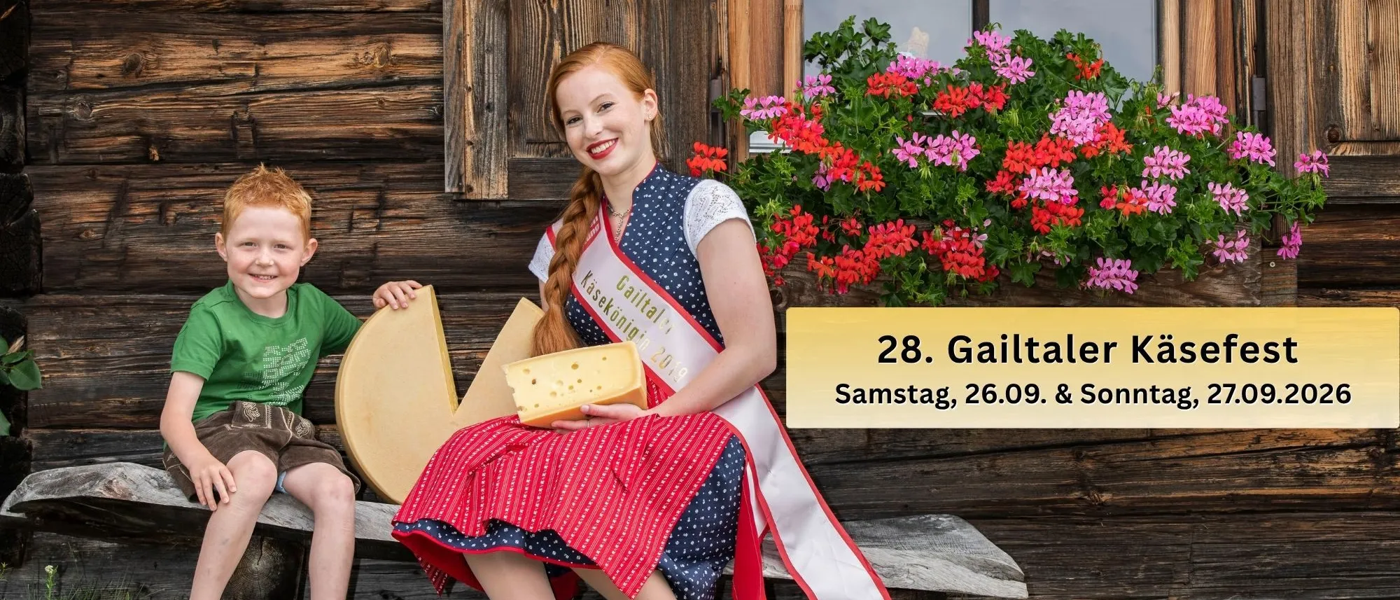 Eine Frau in einem traditionellen Kleid hält ein großes Stück Käse, während ein junger Junge neben ihr auf einer Holzbank vor einem Holzhaus sitzt.