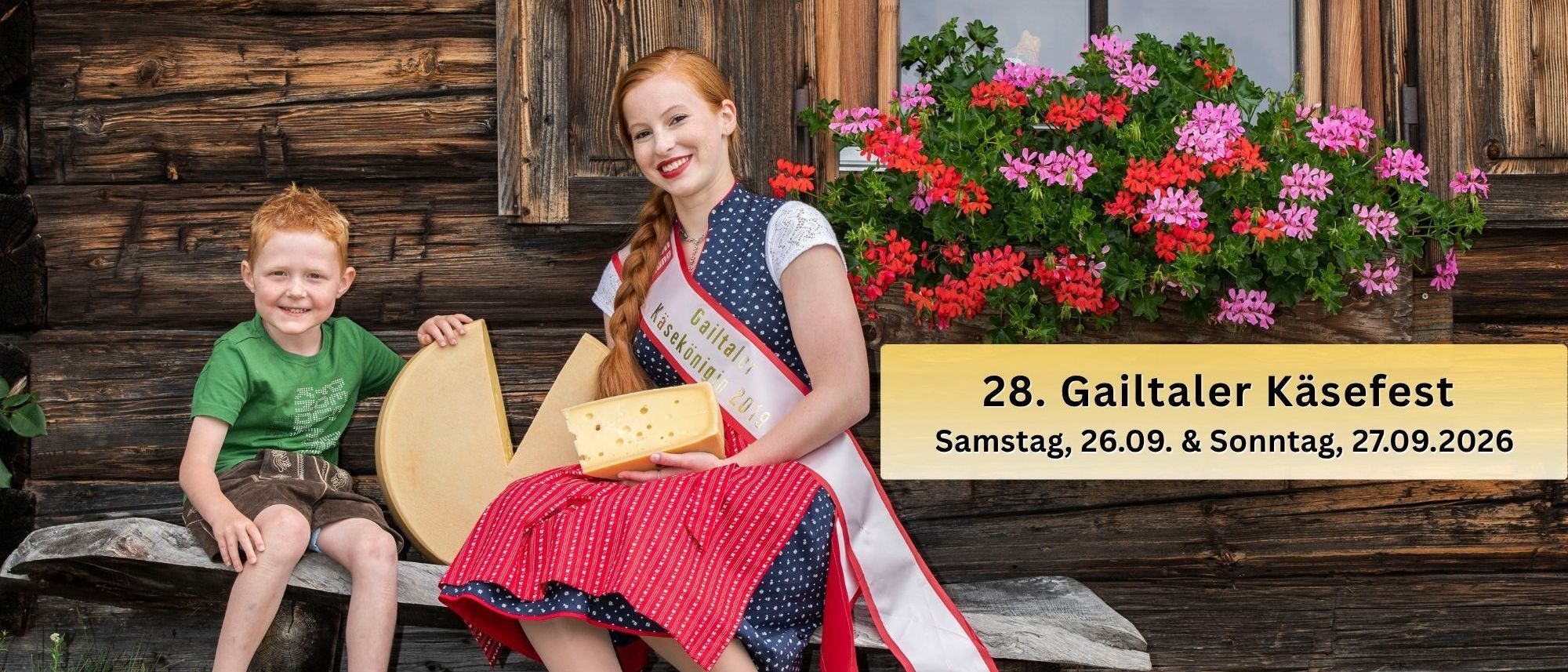 Eine Frau in einem traditionellen Kleid hält ein großes Stück Käse, während ein junger Junge neben ihr auf einer Holzbank vor einem Holzhaus sitzt.