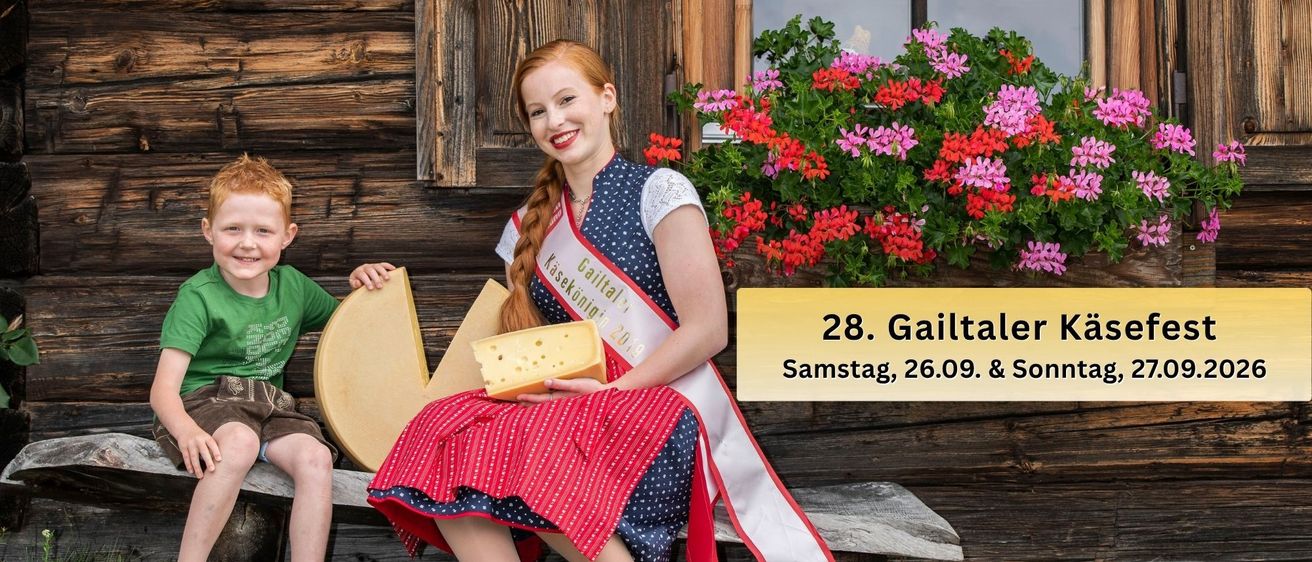 A woman in a traditional dress holds a large piece of cheese while a young boy sits beside her on a wooden bench in front of a wooden house.