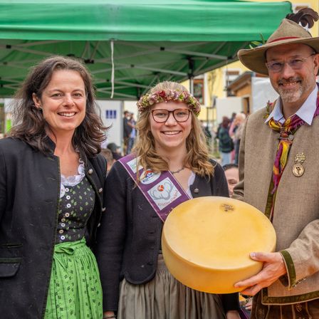 Drei Menschen lächeln unter einem grünen Zelt auf einem Festival. Eine Frau in einem grünen Kleid, eine Frau mit Blumenkranz und ein Mann mit gelbem Käse.