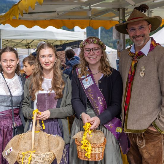 Vier Personen stehen unter einem Zelt und lächeln. Zwei Frauen halten Weidenkörbe, eine trägt eine Krone, die andere eine Schärpe. Ein Mann mit Hut und Brille lächelt neben ihnen.