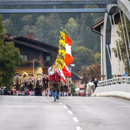 Ein Umzug mit Teilnehmern in traditioneller Kleidung, die eine Straße hinuntermarschieren, eine Fahne mit Löwendesign tragend. Im Hintergrund sind Gebäude, Bäume und eine Brücke zu sehen.