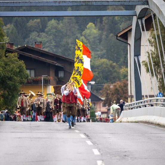 Ein Umzug mit Teilnehmern in traditioneller Kleidung, die eine Straße hinuntermarschieren, eine Fahne mit Löwendesign tragend. Im Hintergrund sind Gebäude, Bäume und eine Brücke zu sehen.