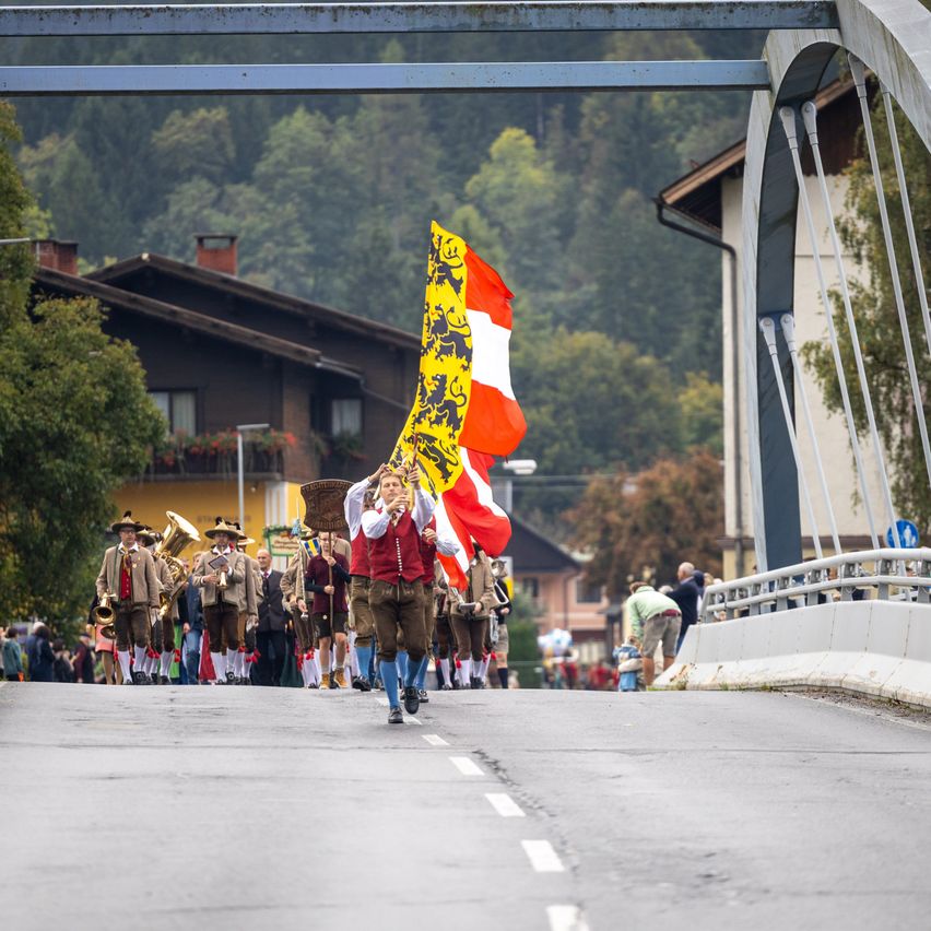 Ein Umzug mit Teilnehmern in traditioneller Kleidung, die eine Straße hinuntermarschieren, eine Fahne mit Löwendesign tragend. Im Hintergrund sind Gebäude, Bäume und eine Brücke zu sehen.