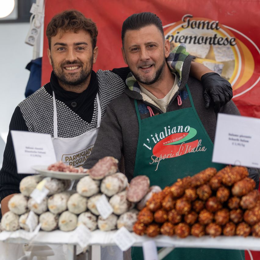 Zwei Männer lächeln und posieren für ein Foto an einem Lebensmittelstand mit verschiedenen Fleischprodukten zur Schau.