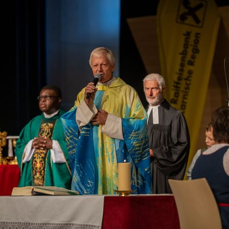 Ein älterer Priester spricht in ein Mikrofon auf einer Bühne. Er trägt eine blau-gelbe Robe. Hinter ihm steht ein weiterer Priester. Ein brennendes Kerze und ein Buch liegen auf dem Tisch vor ihm.