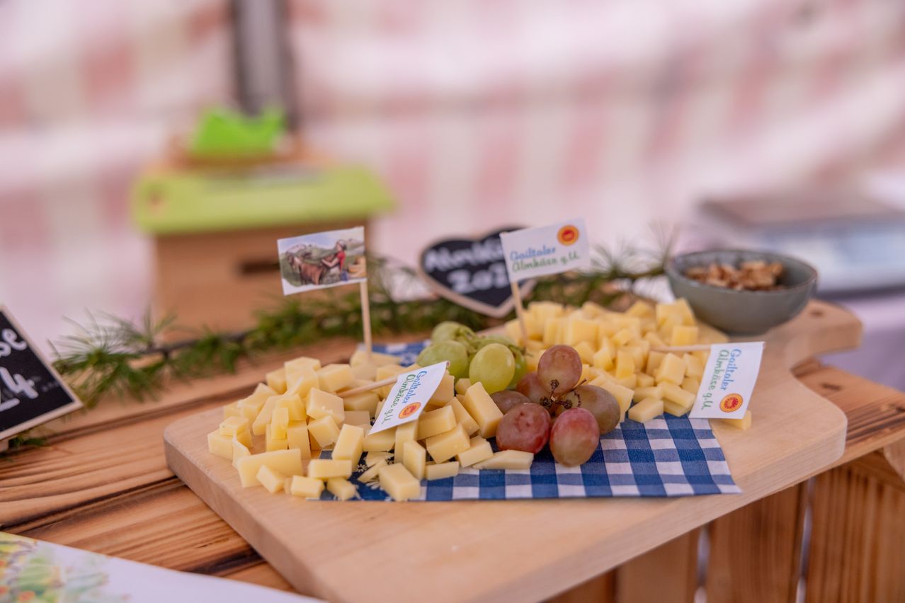 Auf einem Holzschneidebrett befinden sich verschiedene Käsesorten und Trauben. Etiketten zeigen die Käse- und Traubensorten an.