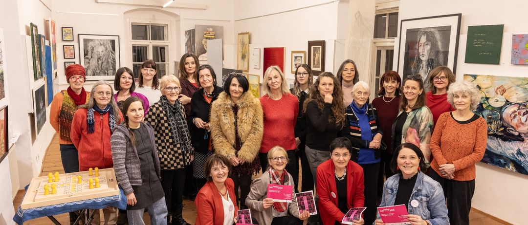 Eine Gruppe von Frauen steht in einem Raum, hält rosa Broschüren und mehrere Gemälde an den Wänden, mit einem Holzboden.