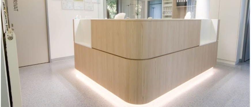 Eine moderne Rezeptionstheke in einem Büro mit Holzpaneelen, Glastrennwänden und beleuchteter Beleuchtung. Die Theke hat eine weiße Oberseite und wird von Leuchten beleuchtet.