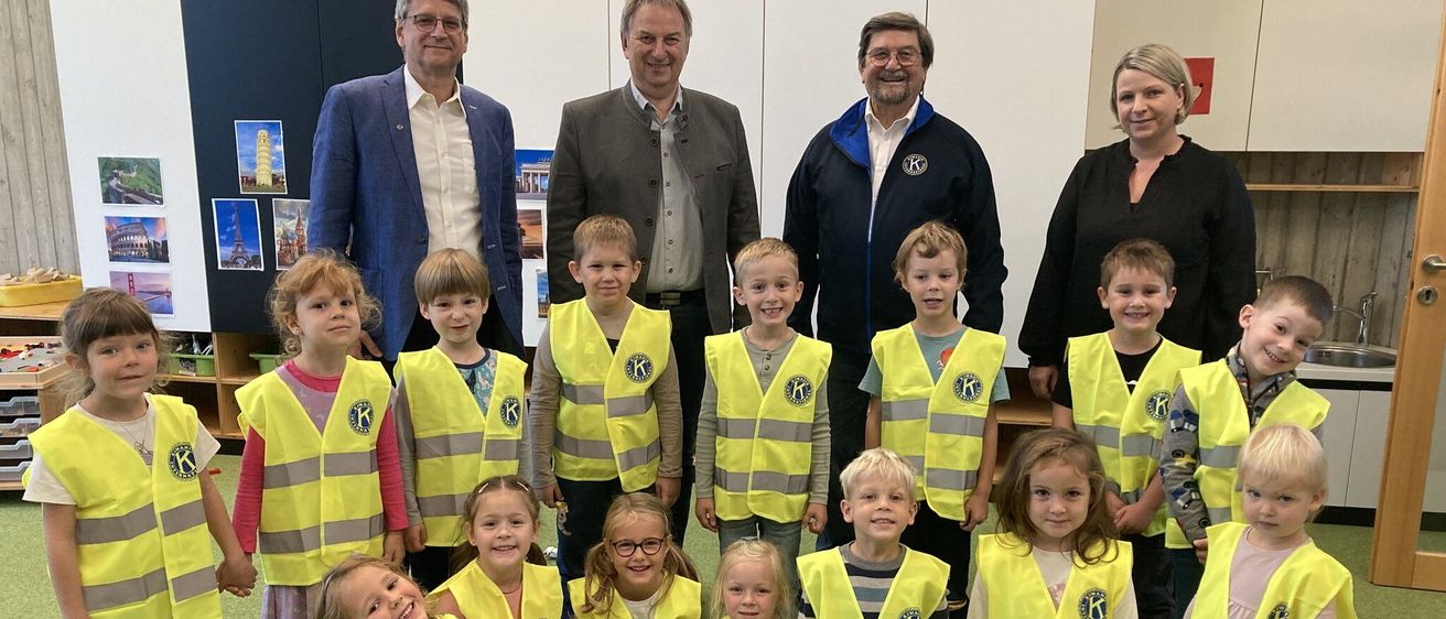Eine Gruppe von Kindern und Erwachsenen, die Sicherheitswesten tragen, posiert für ein Foto in einem Klassenzimmer. Die Kinder sitzen auf dem Boden, während die Erwachsenen hinter ihnen stehen.