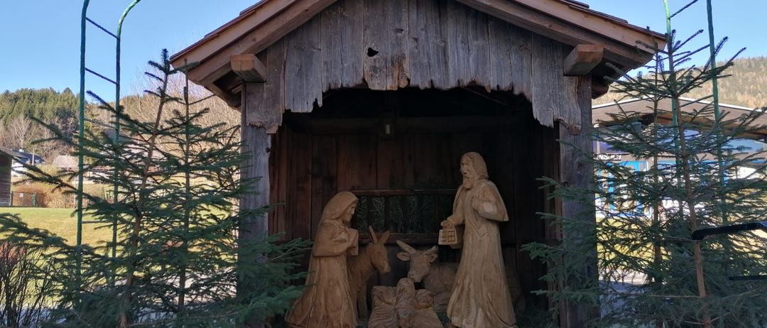 Eine hölzerne Krippe mit Josef, Maria, Jesus und Tieren unter einem Holzdach mit einem grünen Bogen im Hintergrund.