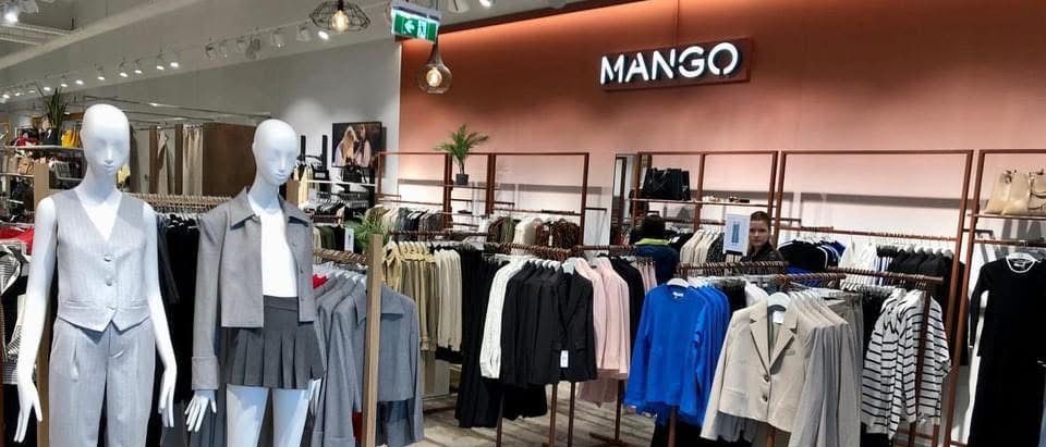 Ein Bekleidungsgeschäft namens Mango mit Mannequins in Röcken und Jacken, sowie Kleidung an Kleiderständern, einem Topf auf einem Regal und einem Mitarbeiter im Hintergrund.